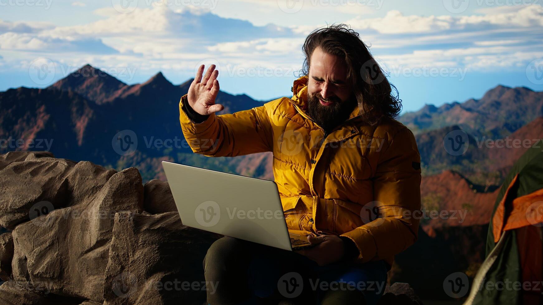 Mountain adventurer prioritizes his online business over safety, working on his laptop despite the extreme winds. The breathtaking landscape is overshadowed by his internet dependency. Camera A. photo