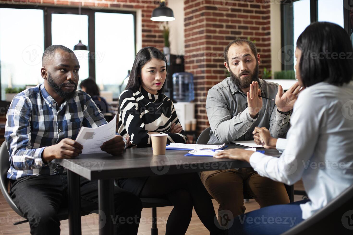Black female applicant sits in a formal interview with diverse recruiters at office desk. Asian woman and male colleagues ask serious questions, evaluating the job candidate in brick wall workplace. photo