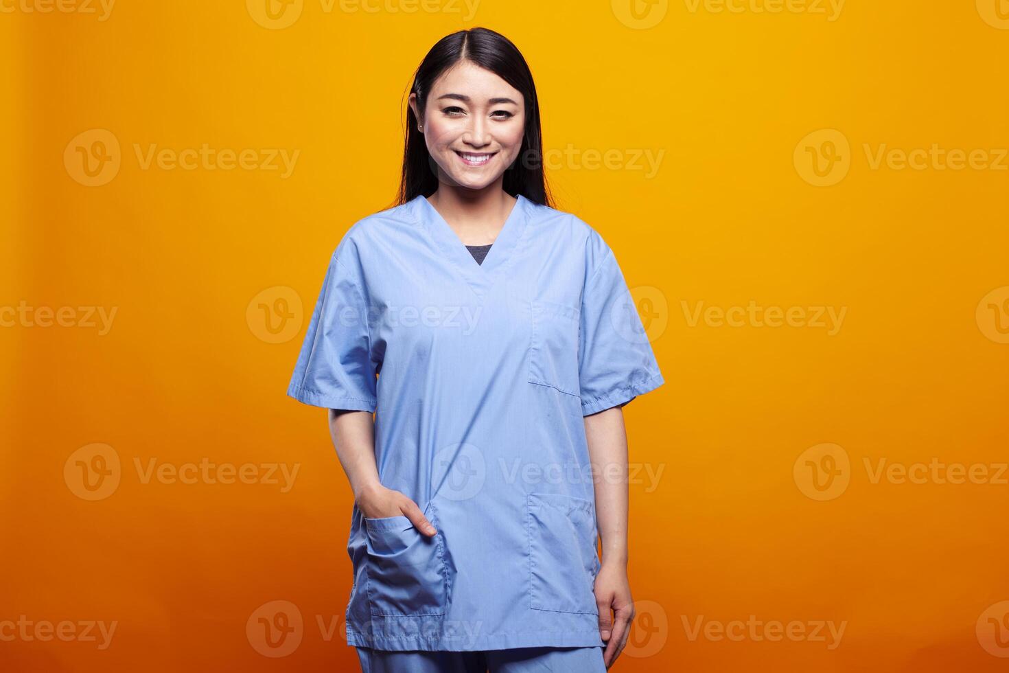 Young woman in medical scrubs, having hand in a pocket and posing in front of yellow background. Portrait of female asian nurse standing and smiling at camera, portraying readiness for healthcare. photo