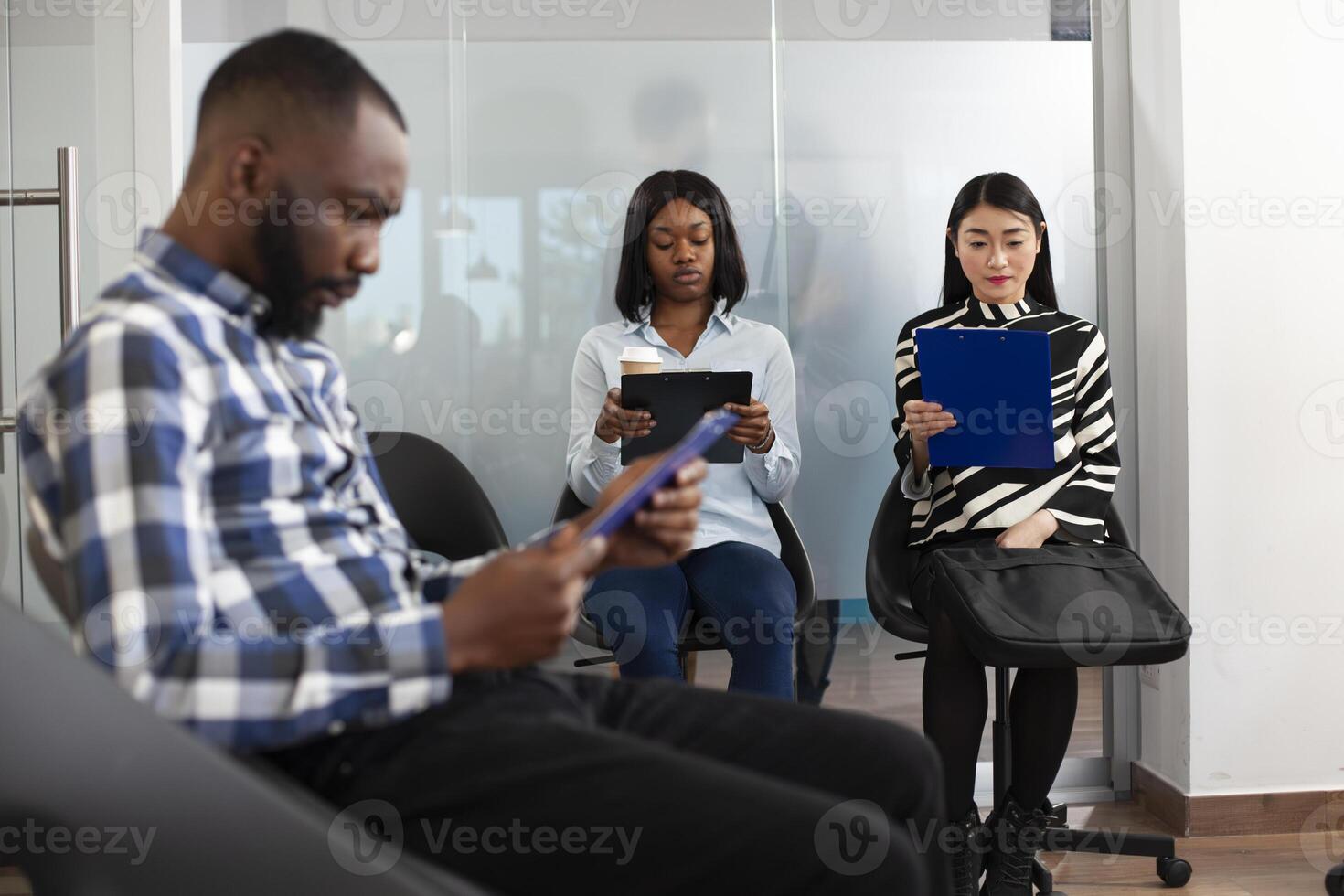 Young women seated together in office lobby, reading company regulations and waiting for interview. Multiethnic male and female job applicants sitting in queue and preparing for employment meeting. photo