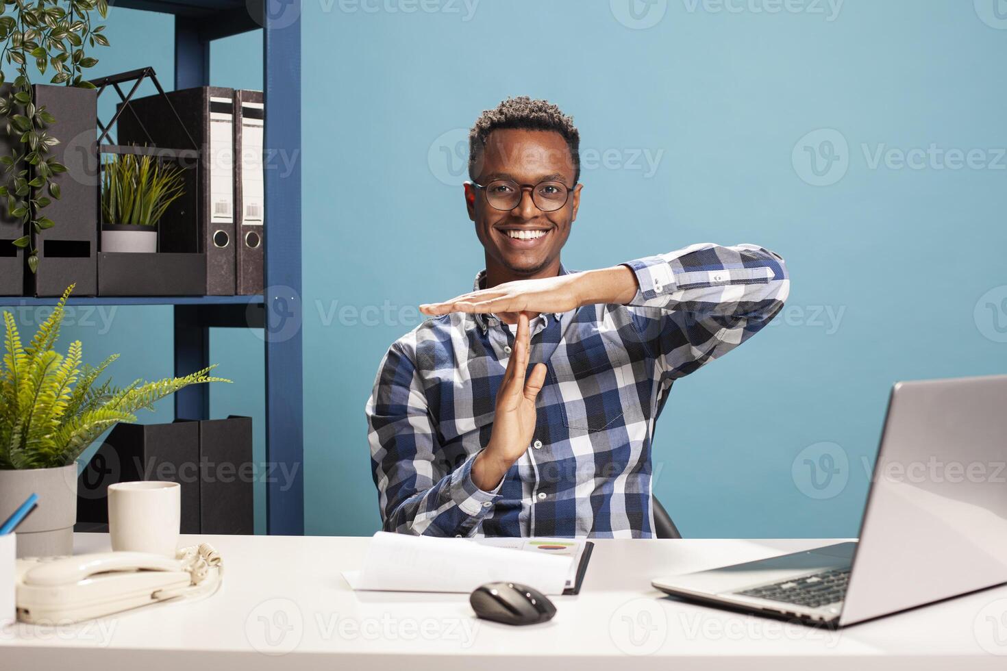 Black male researcher sitting at his desk with a smile is doing a time out gesture towards the camera. Portrait of arican american man making a pause signal using his hands on a blue background. photo