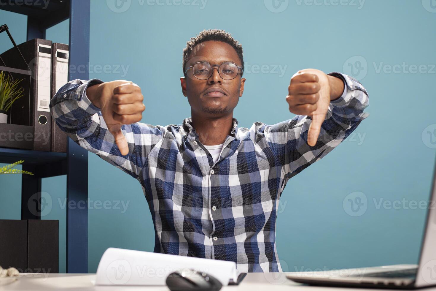 Closeup of black man giving two thumbs down towards camera, expressing disapproval. African american male individual sitting at his desk and gesturing rejection with his hands signaling downwards. photo