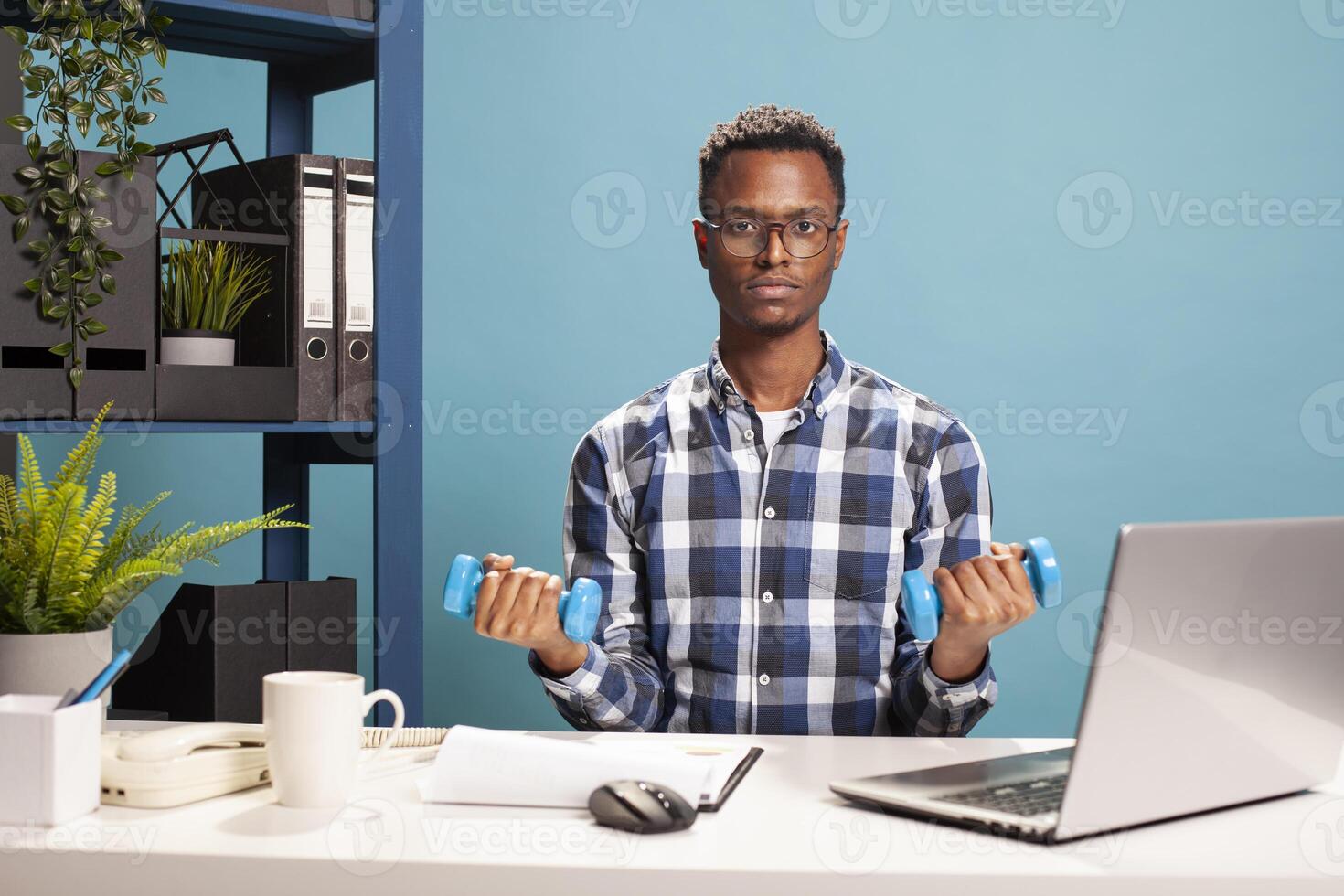 African american businessman seated at office desk, doing arms workout with dumbbells. Active young professional maintaining fitness, exercising with hand weights during office hours. photo