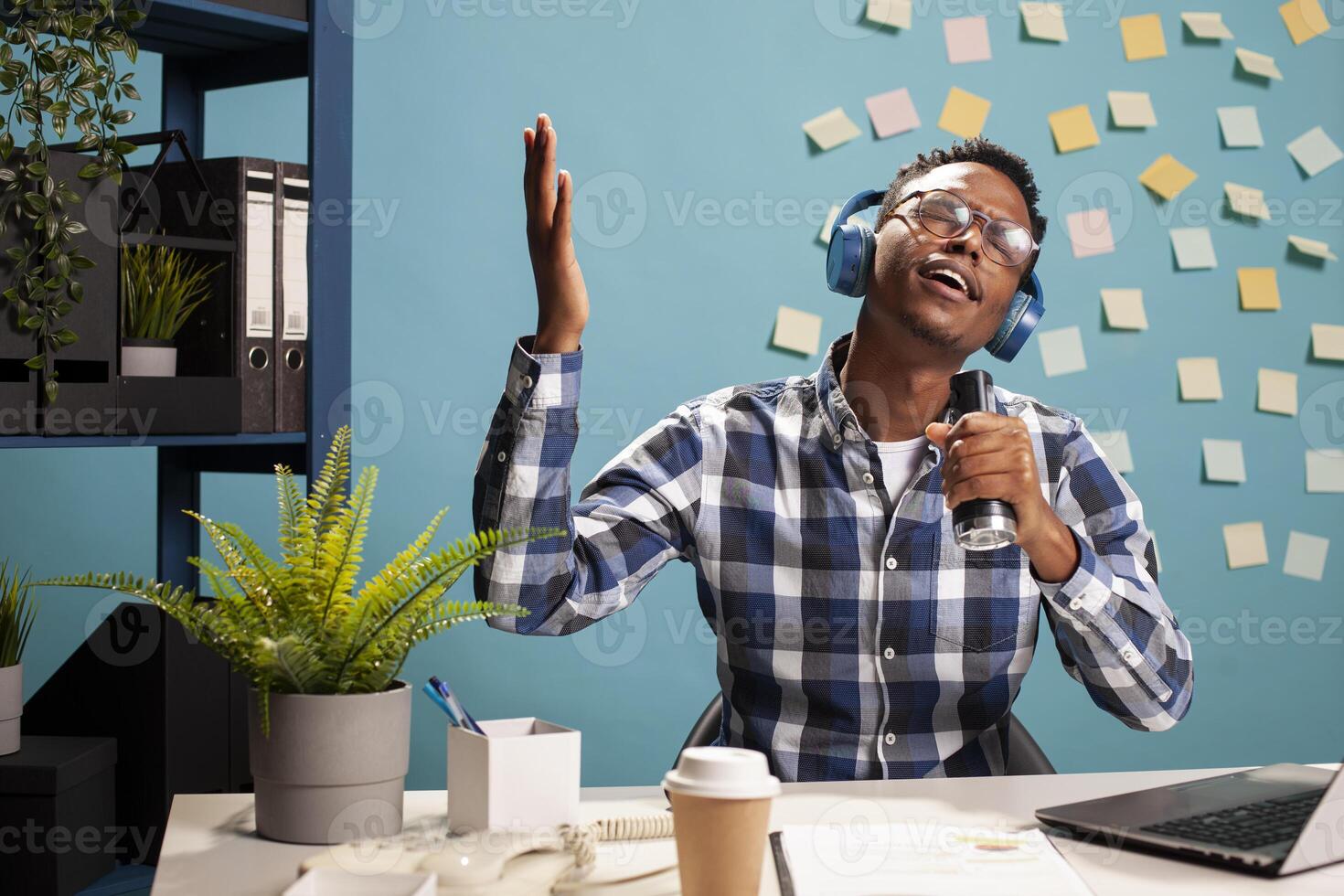 Young project manager wearing wireless headphones, singing along, enjoying music during a break in company workspace. Happy male analyst hand raised, seated at desk with a flashlight, doing karaoke. photo
