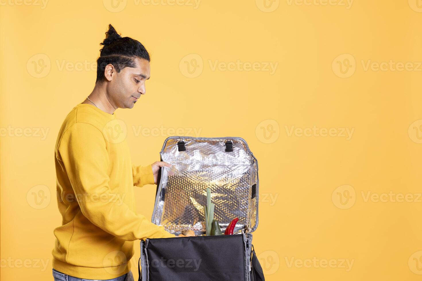 Delivery carrier employee taking takeaway order out of bag, providing it to client. Indian man taking groceries order out of thermal backpack, ordered by customer, studio background photo