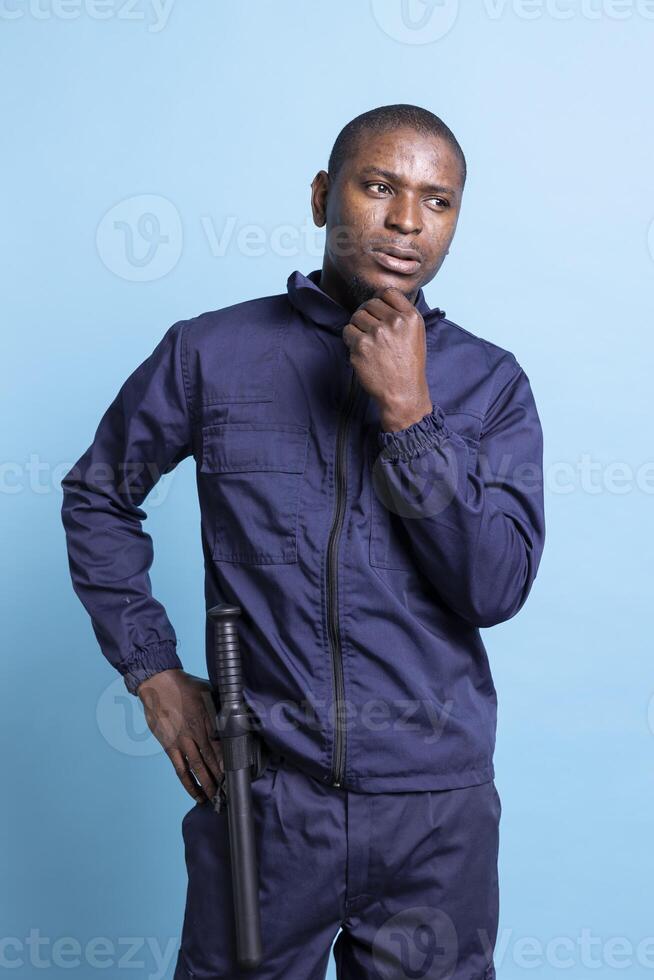 Pensive african american bodyguard thinking about his duty, working on protecting and safeguarding the premises. Male security agent dressed in uniform, armed defense forces in studio. photo