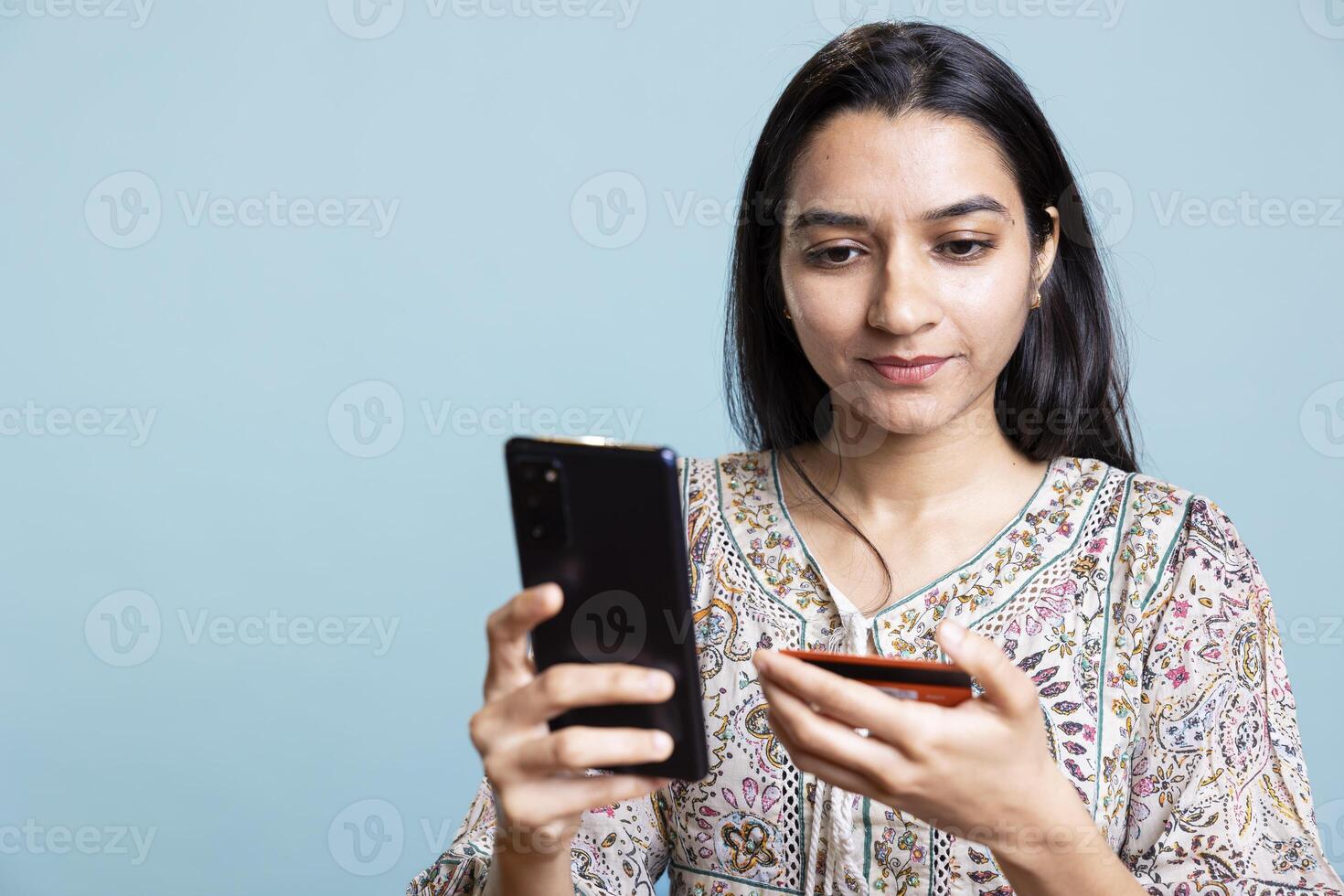 Jolly woman adding payment method on shopping website using phone, taking advantage of promotions in the studio. Indian girl buying stuff online using credit card, typing data on device. photo