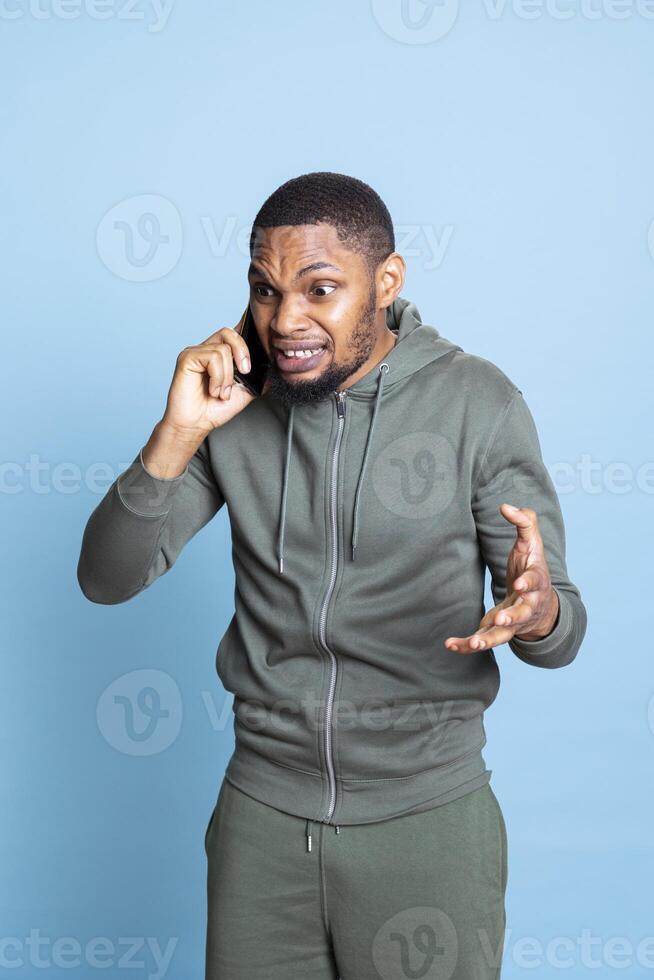 Irked african american man fighting with friends during telephone call, feeling angry and frustrated in studio. Outraged person arguing with pals during conversation on mobile phone. photo