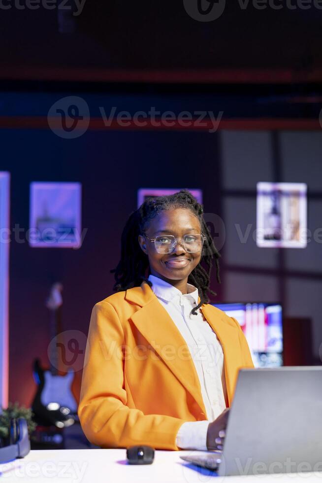 Portrait of jolly woman sitting on chair at home office desk in neon illuminated apartment late at night. Cheerful african american person in RGB lit living room enjoying leisure time photo