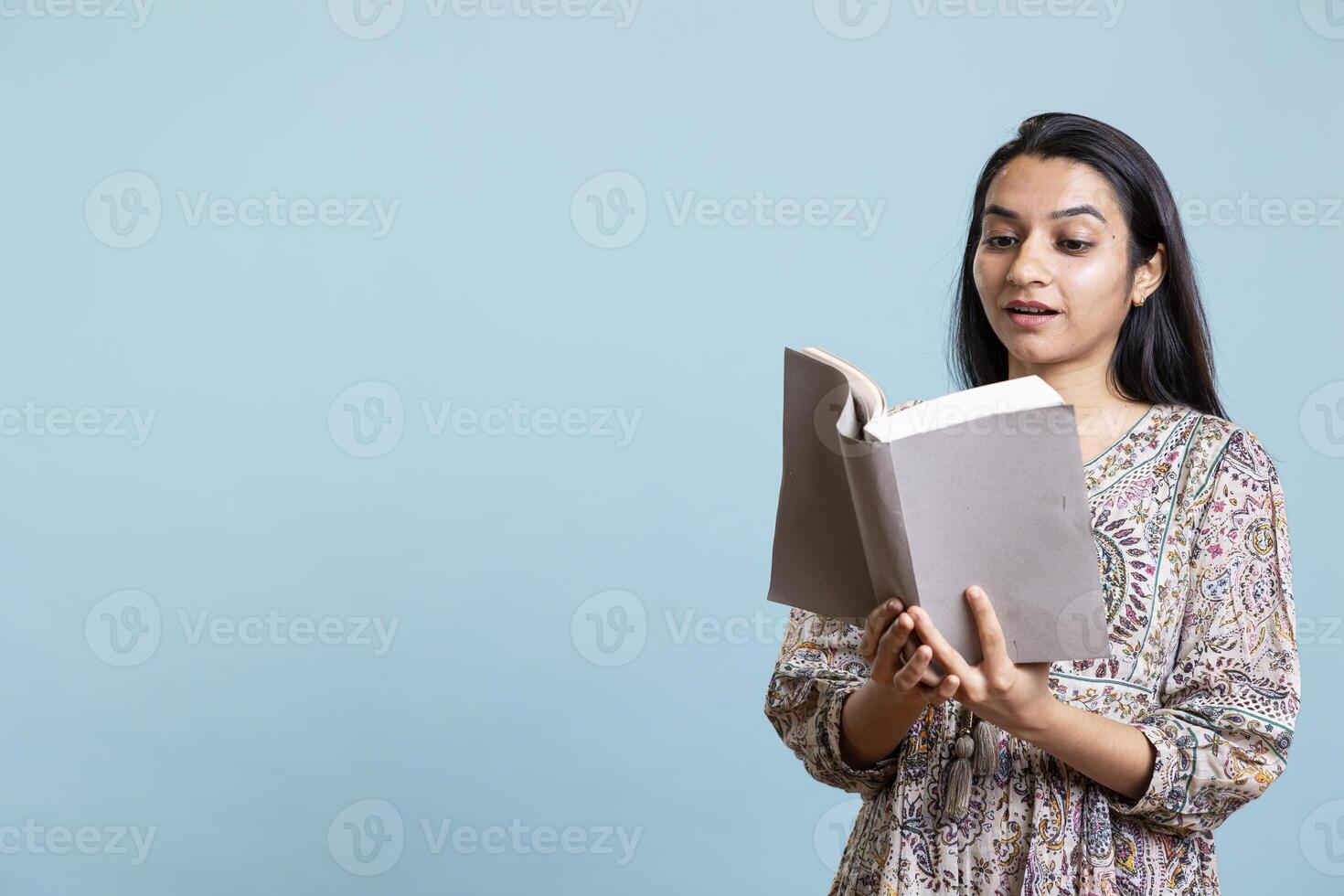 Positive jolly woman reading interesting book and enjoying lecture hobby, being entertained against studio background. Indian relaxed person with intriguing novel in arms enjoys free time. photo