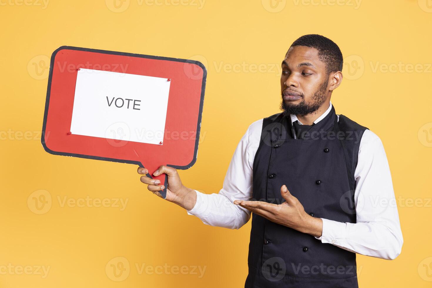 Catering employee showing a message to vote on red speech bubble, presenting the cardboard icon to do a commercial for politics. Waiter in uniform works on customer service. photo