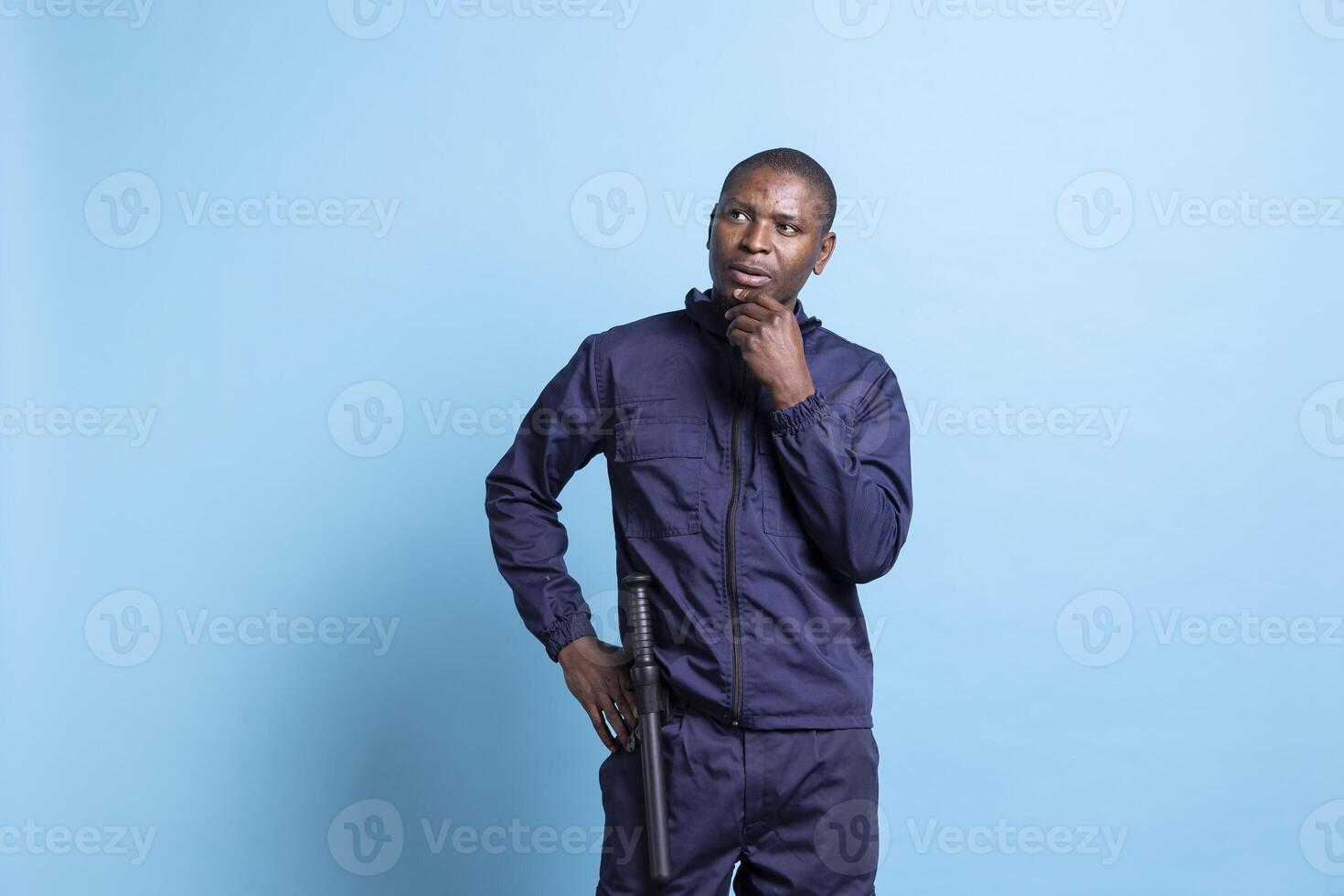 Thoughtful security agent thinking of patrol duties in studio, dressed in guarding uniform with baton. African american safety agent contemplates on controlling access to the premises. photo
