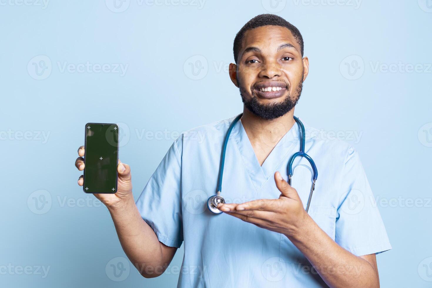 Portrait of joyful african american nurse presenting isolated mockup screen on phone. Smiling healthcare staff pointing at his smartphone with copy space display, studio backdrop. photo