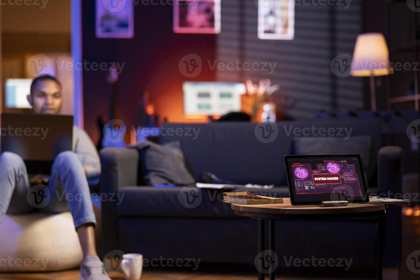 Black man sitting on a bean bag coding on his laptop for cyber security. Tablet warns of a cyber attack, reflecting online security risks, data theft, malware and unauthorized access concerns. photo