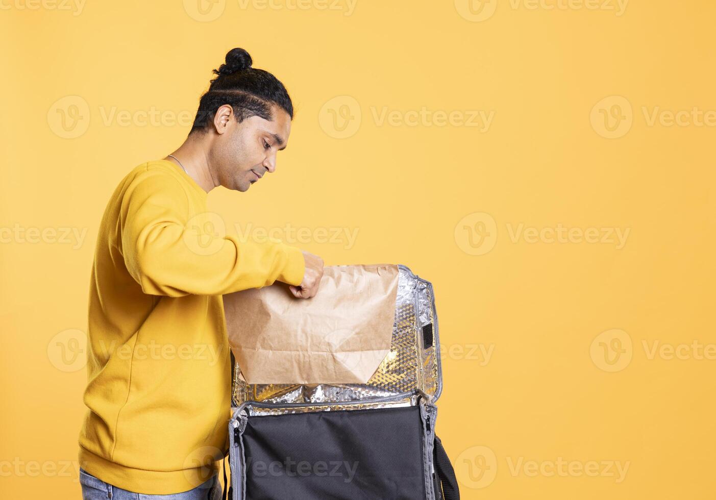 Delivery man placing paper bag with takeaway order in thermal backpack. Food courier putting packaged meal in transportation bag, preparing to go to client address, isolated over studio background photo