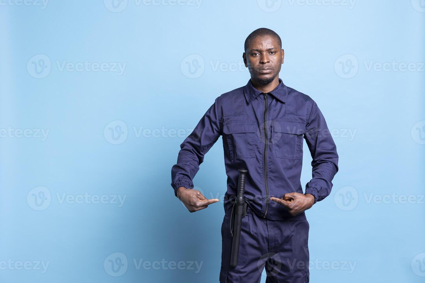 Armed security officer presenting his baton weapon for own safety in studio, pointing at his police bat to ensure order and protection. Male trained personnel in uniform against blue background. photo