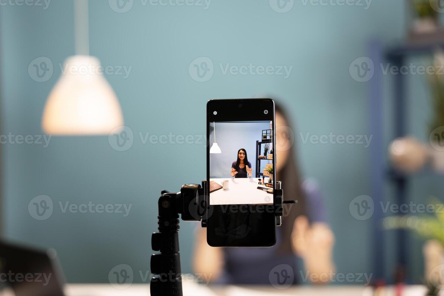 A smartphone camera captures a beauty influencer reviewing cosmetics. Mobile device screen showing asian vlogger using expressive gestures as she explains beauty products in a blue background studio. photo