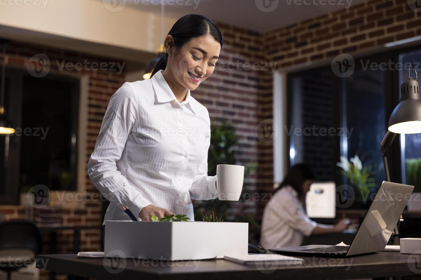 Smiling female manager stands and arranges her desk, enjoying the process of decorating her office. She holds a coffee cup in one hand, as she delights in the organization of her workspace photo