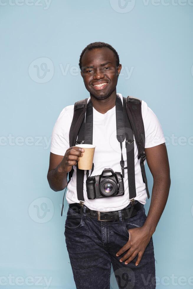 Portrait of a smiling photographer with a camera around his neck, standing and holding a warm beverage. Cheerful black man capturing professional headshots for his portfolio, with coffee in his hand. photo
