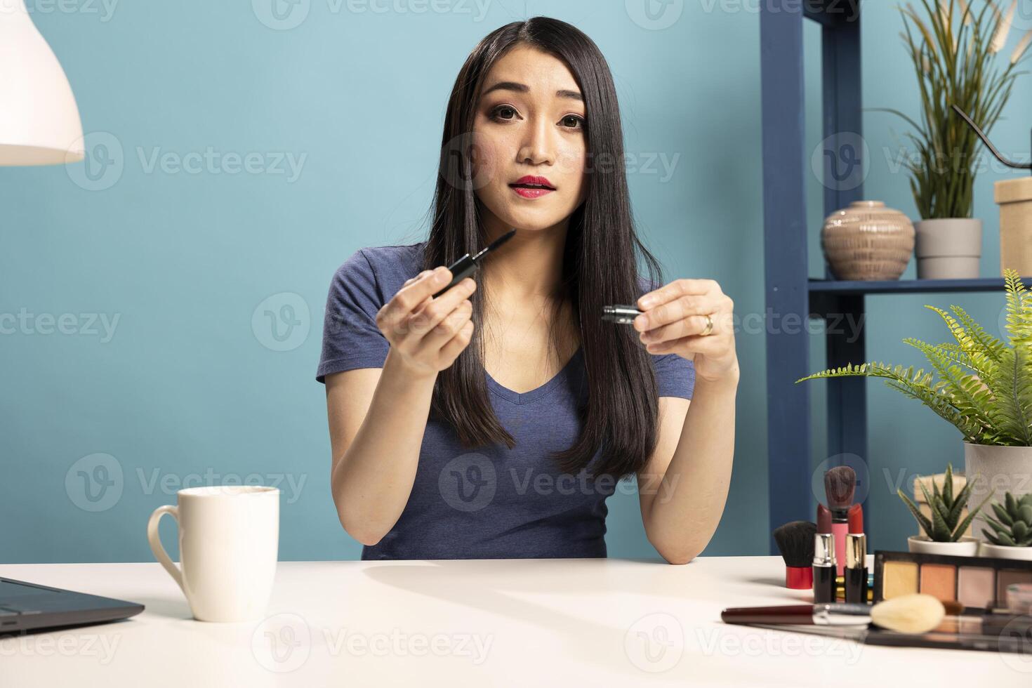 Young makeup artist holding a mascara, filming a tutorial for her online viewers. Asian female vlogger sitting at desk, recording livestream to showcase her beauty routine in blue background studio. photo