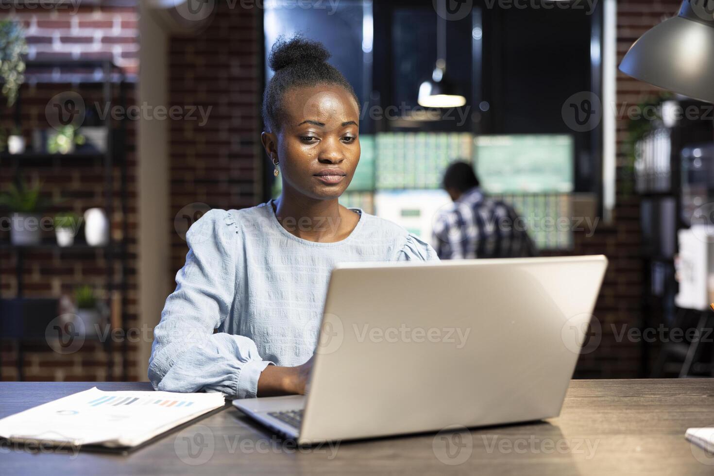 Determined female entrepreneur analyzing financial data on laptop at her desk, surrounded by trading documents. Her coworker, slightly blurred, works in the background at a multiscreen setup. photo