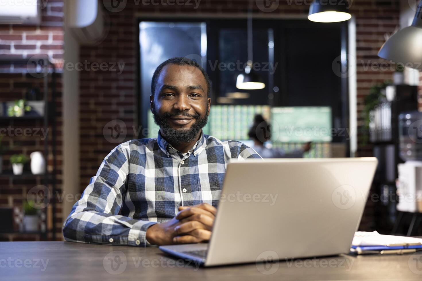 African american male professional sitting at a desk in a modern office with a brick wall, looking at the camera and smiling. His laptop and clipboard are set up for reviewing financial data. photo