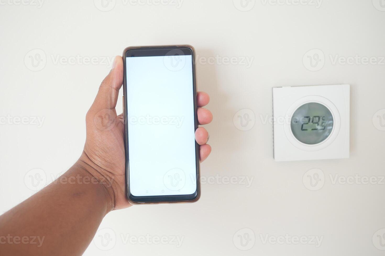 Hand holding smartphone next to digital thermostat showing temperature photo