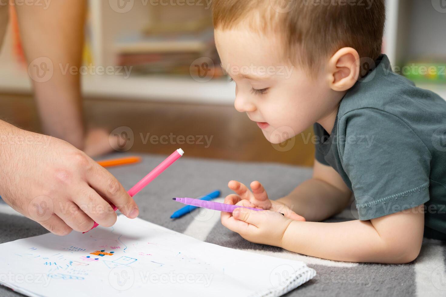 A beautiful little toddler boy of two or three years old with dad draws with markers in the album in the children's room at home sitting on the floor. Spending time with children. Selective photo