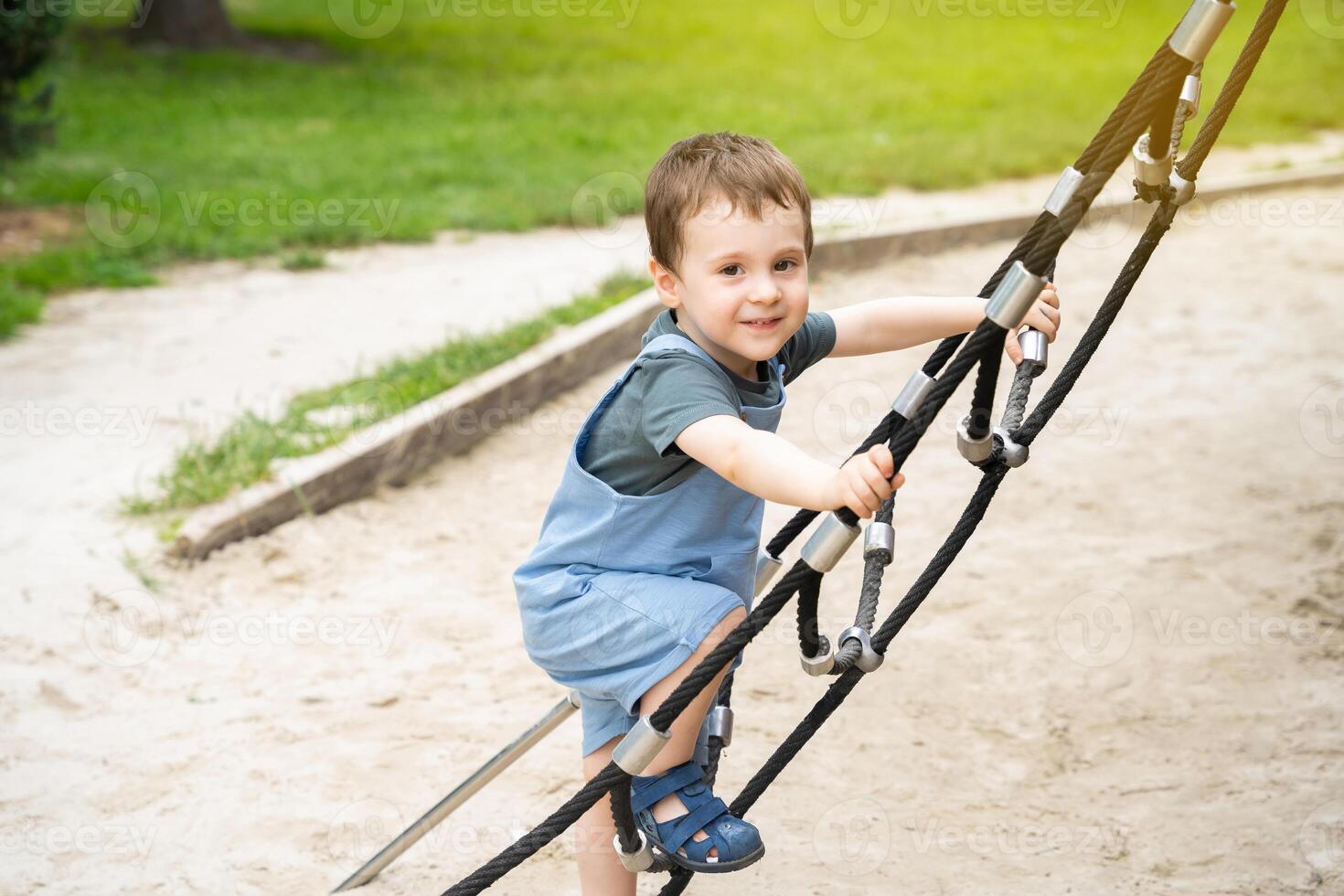 Happy toddler boy 2.5 years old climbs the rope ladder at the playground in the park. Active games and entertainment for children. sunny summer day photo