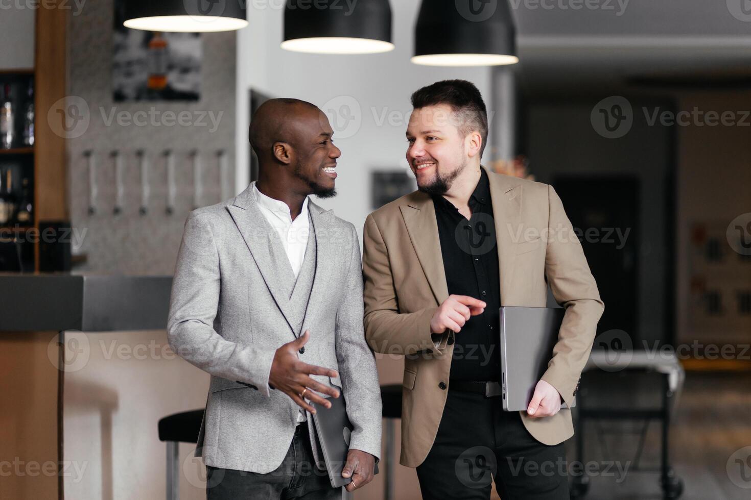 Smiling excited young African American businessman discussing project over business lunch. Two happy professionals men discussing financial market data standing at corporate office hall photo