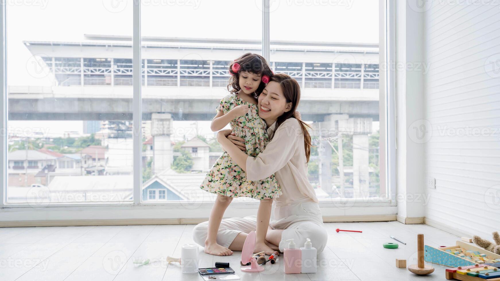Happy mother hugging her daughter lovingly while sitting in a bright living room, surrounded by toys and makeup during a playful bonding moment at home. photo