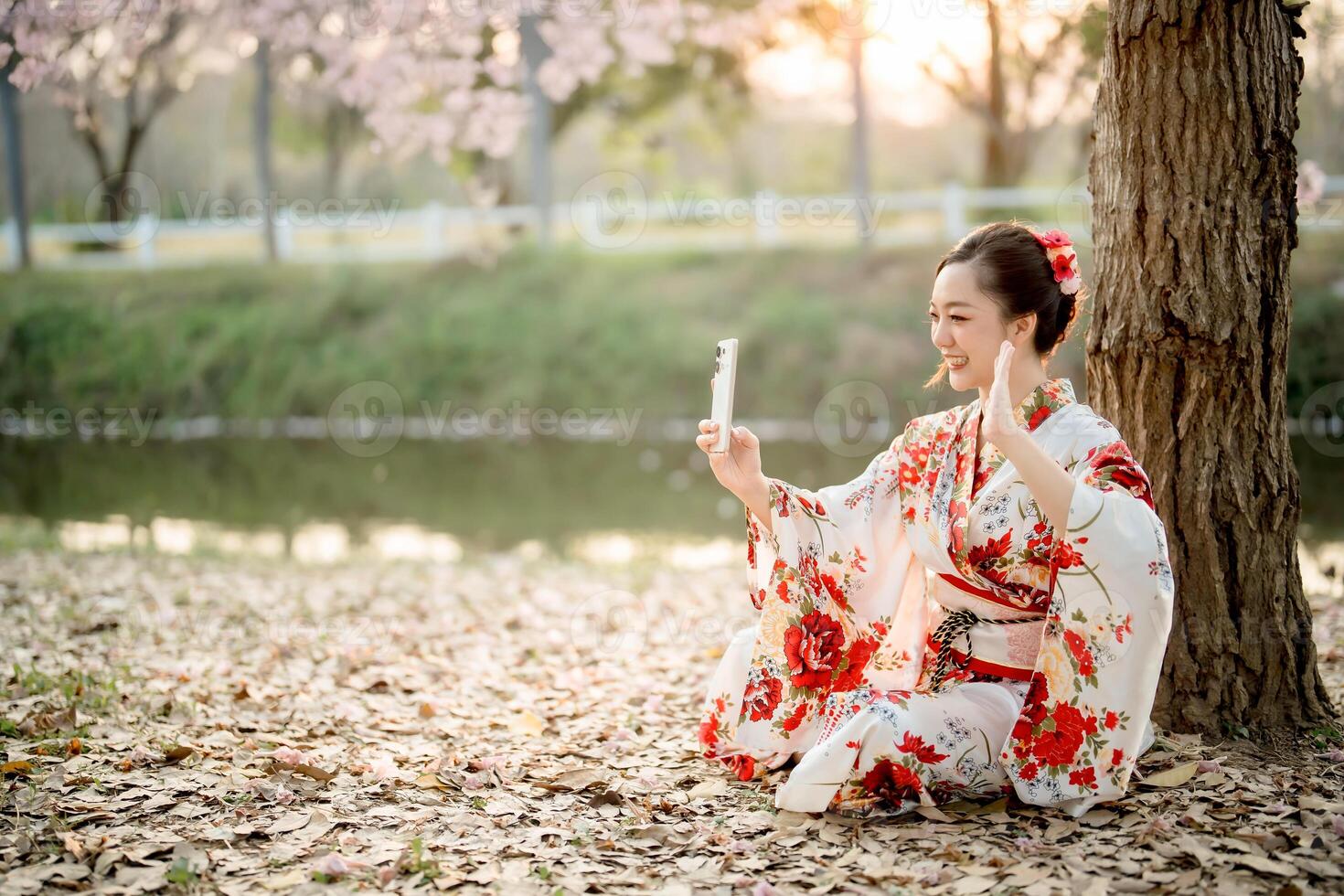 Smiling Asian woman in traditional floral kimono chatting on smartphone under tree by the river during sunset, blending heritage with modern lifestyle photo