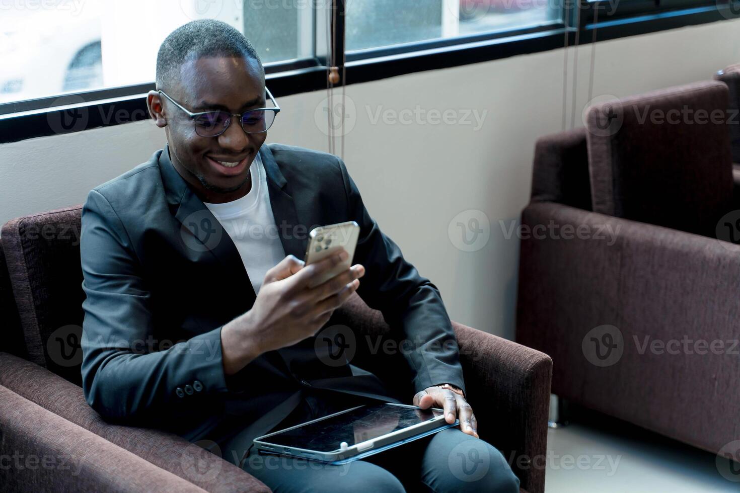 Smiling businessman using smartphone while sitting in modern lounge with tablet on lap, relaxed and engaged in communication or social media browsing photo