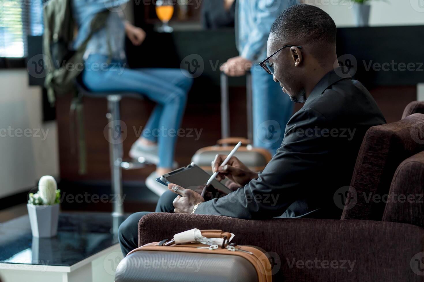 Businessman using digital tablet with stylus while waiting in airport lounge, surrounded by travel luggage and casually dressed travelers in the background photo