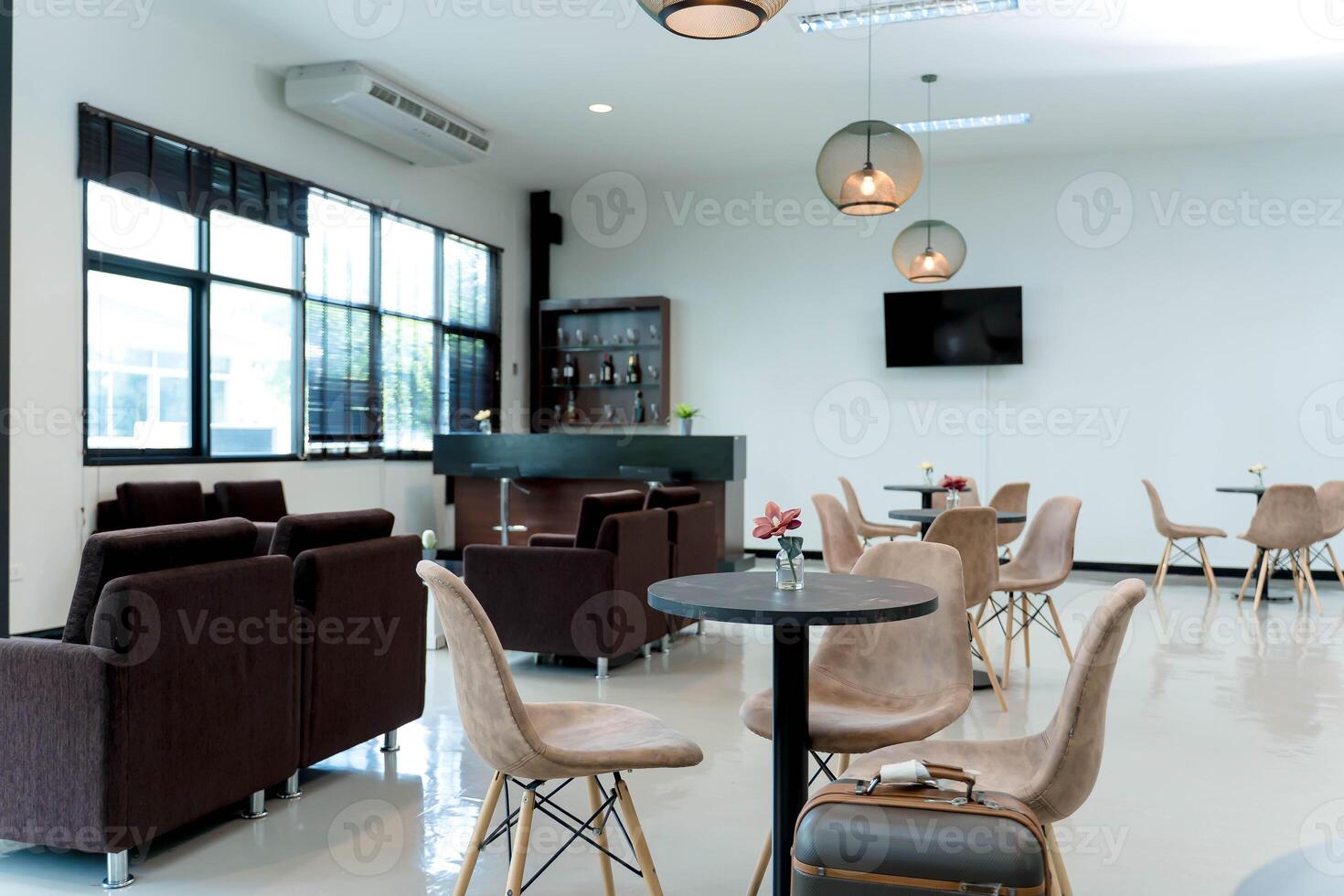 Modern lounge interior with beige chairs, black round tables, small flower vases, and travel suitcase in a bright waiting area with a bar counter photo