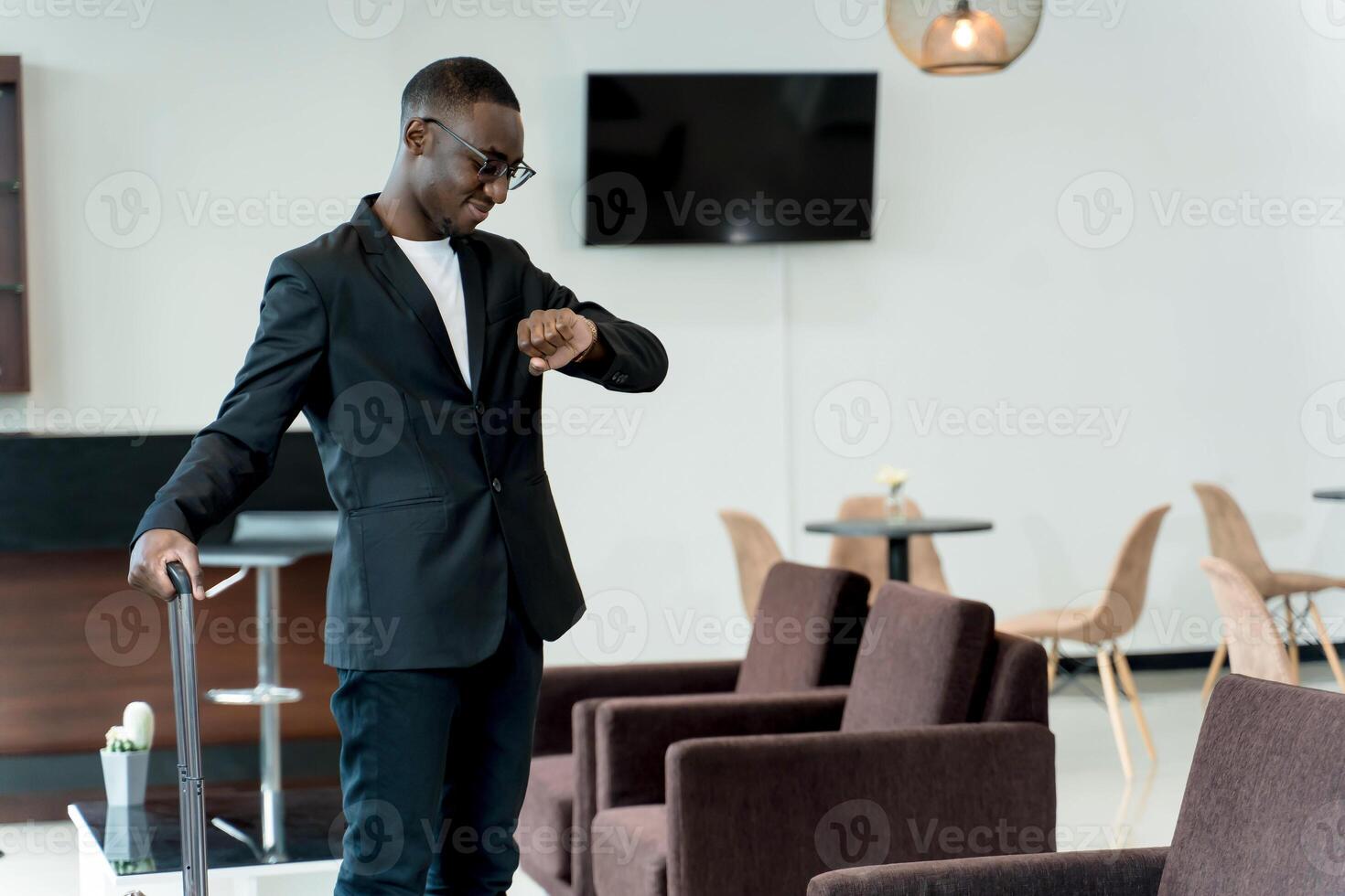 Stylish businessman with suitcase checking the time on his watch while waiting in a modern airport lounge, ready for departure or business travel photo
