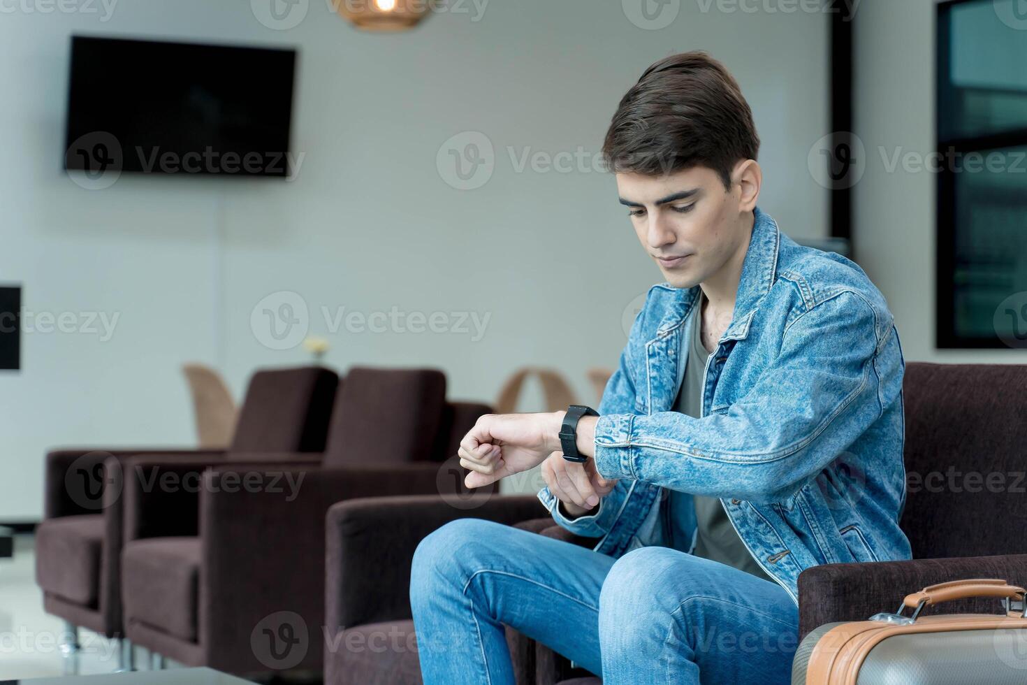 Young male traveler checking time on smartwatch while waiting at modern airport lounge with suitcase, dressed in denim, preparing for departure photo