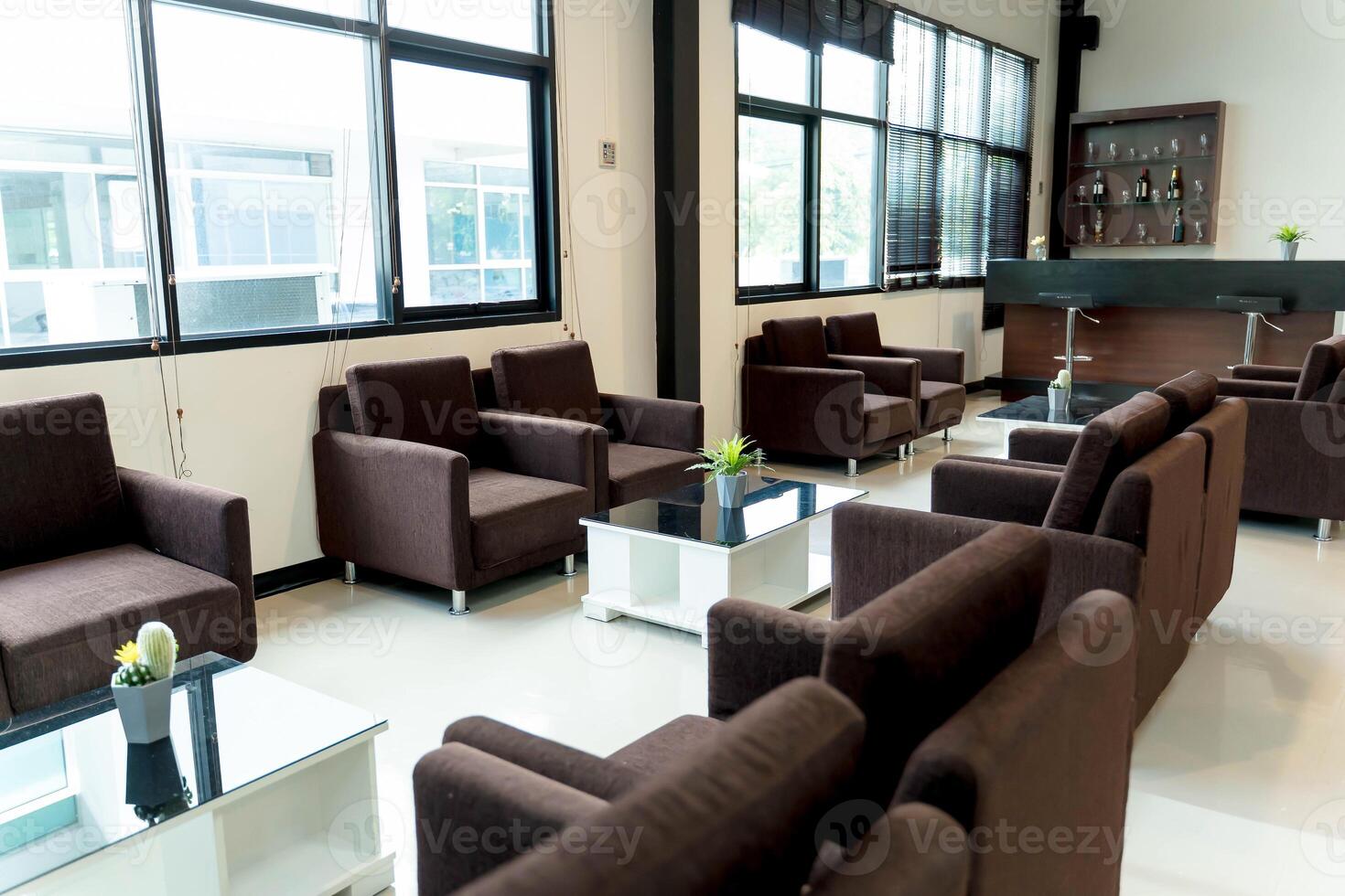 Modern lounge interior with brown fabric armchairs, glass coffee tables, and bar counter in bright room with large windows and minimalist decor photo