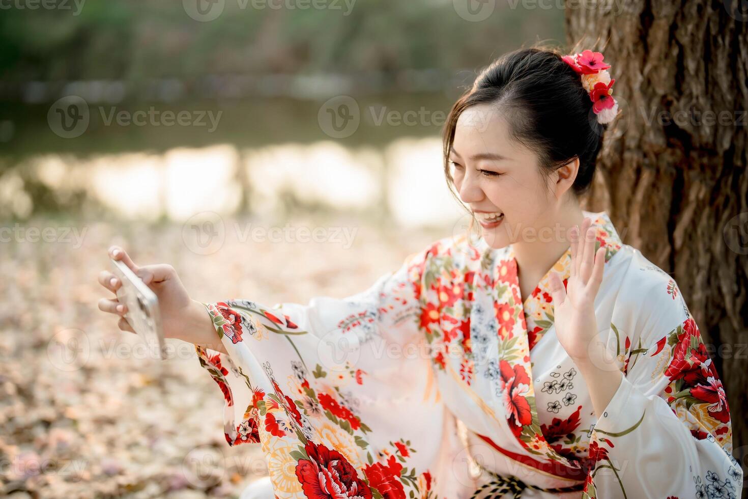 Smiling Asian woman in traditional floral kimono waving during call with smartphone under tree in natural setting, joyful moment with digital connection photo