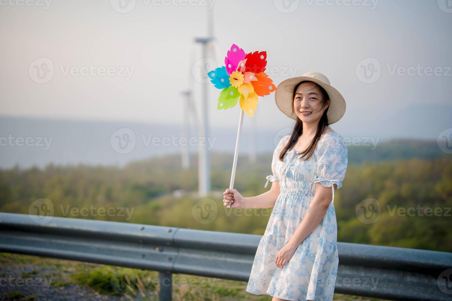 Smiling woman holding a colorful flower-shaped pinwheel, standing outdoors in a summer dress and sunhat near wind turbines with a scenic landscape background photo