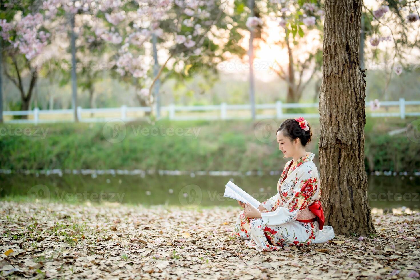 Asian woman in traditional floral kimono reading book peacefully under tree by riverside during spring sunset, enjoying relaxing moment in nature and culture photo