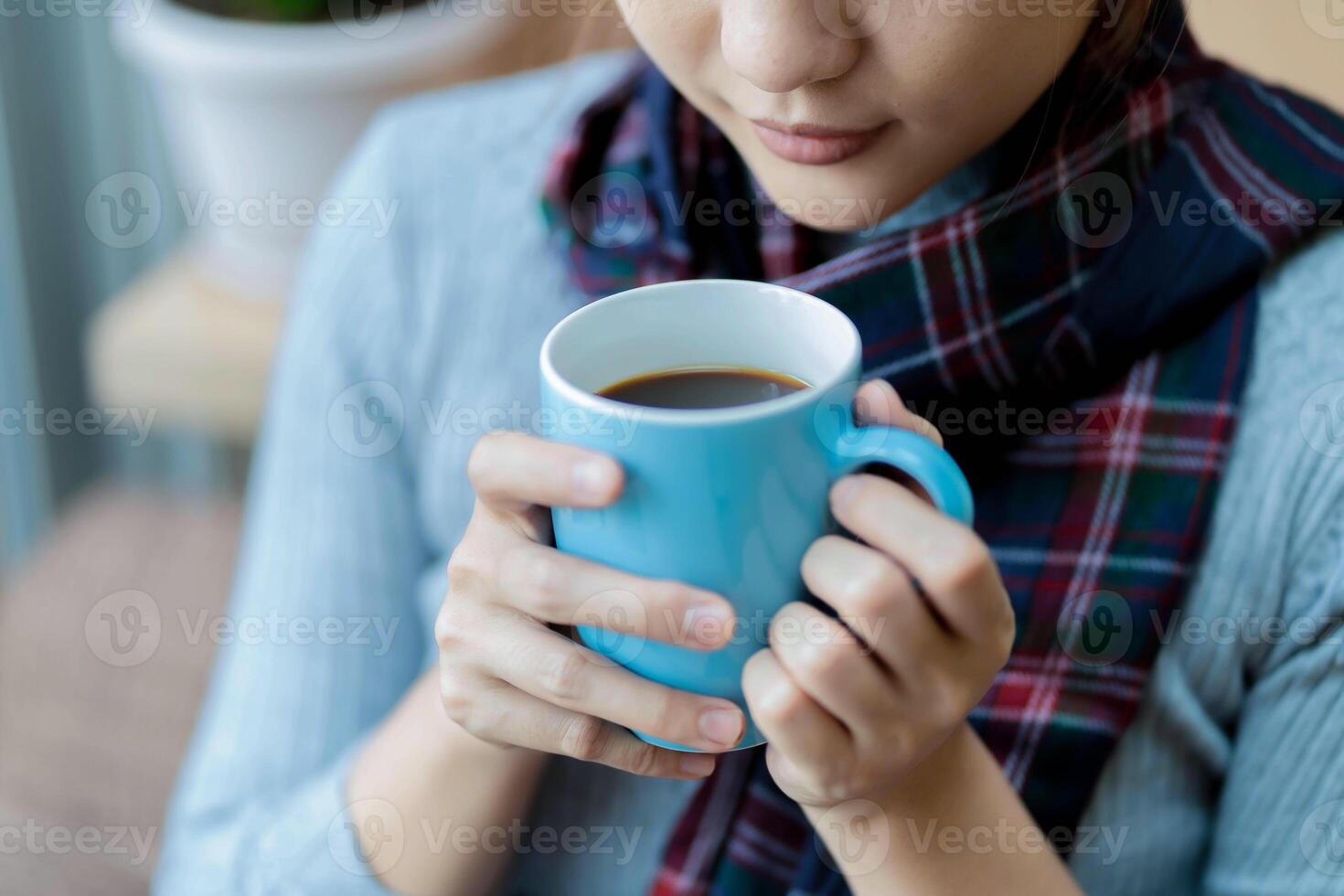 Close-up of woman gently holding a blue coffee mug while wearing a cozy plaid scarf, enjoying a moment of calm and warmth in a relaxed indoor environment photo
