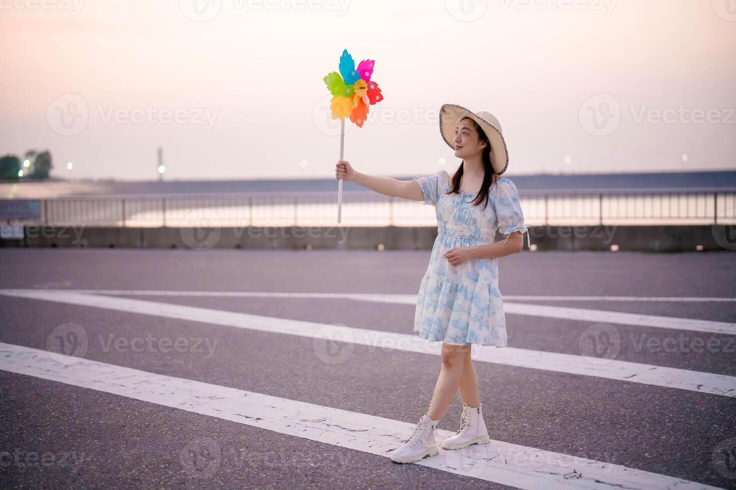 Young woman in a summer dress and sun hat holding colorful pinwheel while walking across a wide road at sunset, enjoying freedom and peaceful outdoor vibes. photo