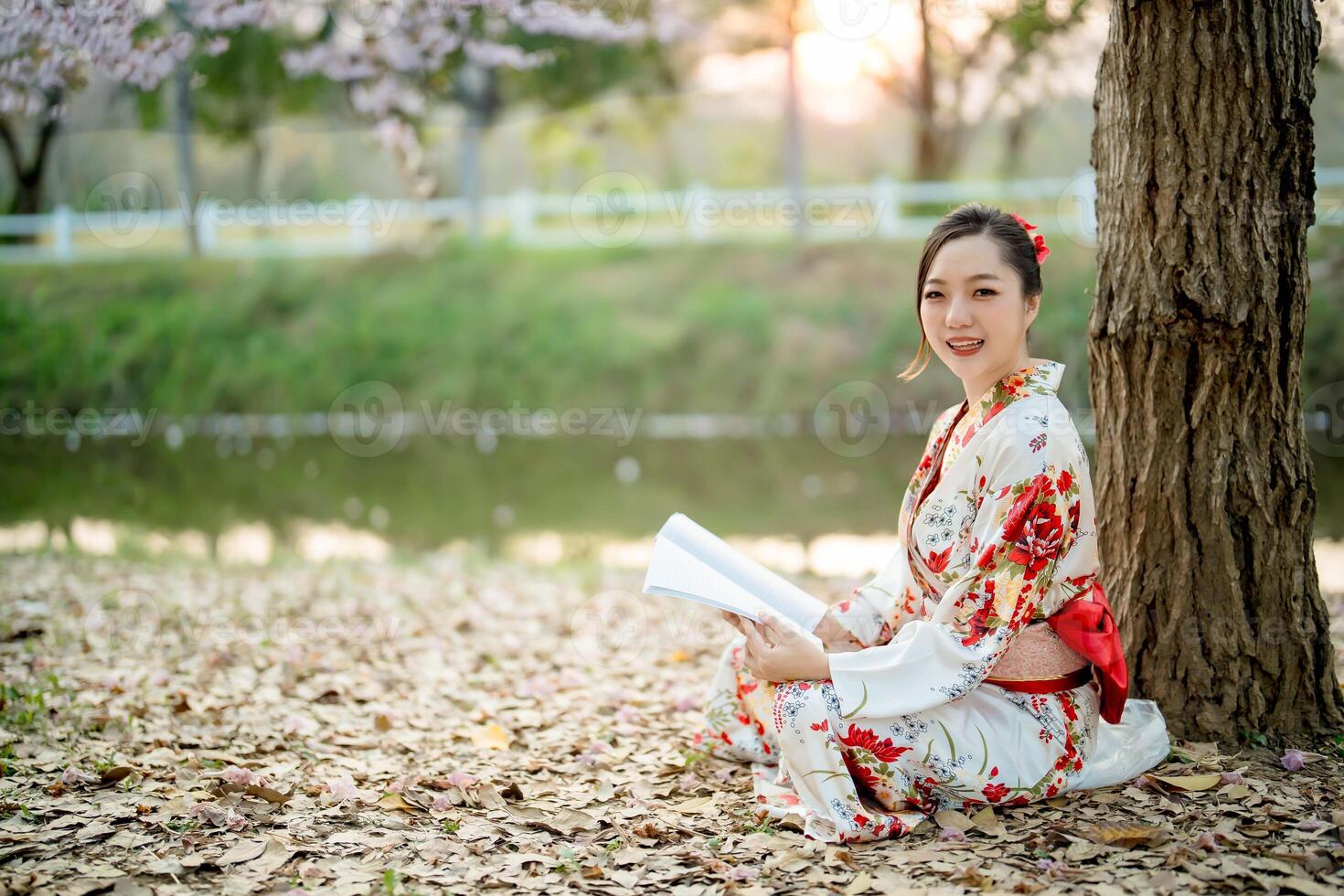 Smiling Asian woman in traditional floral kimono reading book while sitting under tree by the river during peaceful spring sunset in natural outdoor setting photo