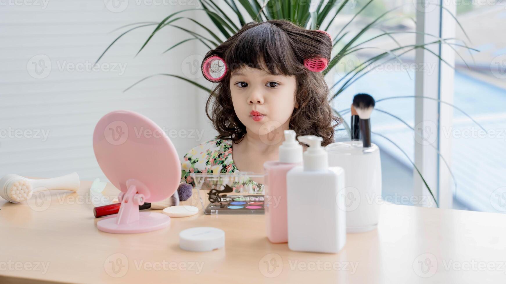 Cute little girl with hair curlers playing with makeup at vanity table, making a kissy face in front of a mirror, imitating adult beauty routine playfully. photo