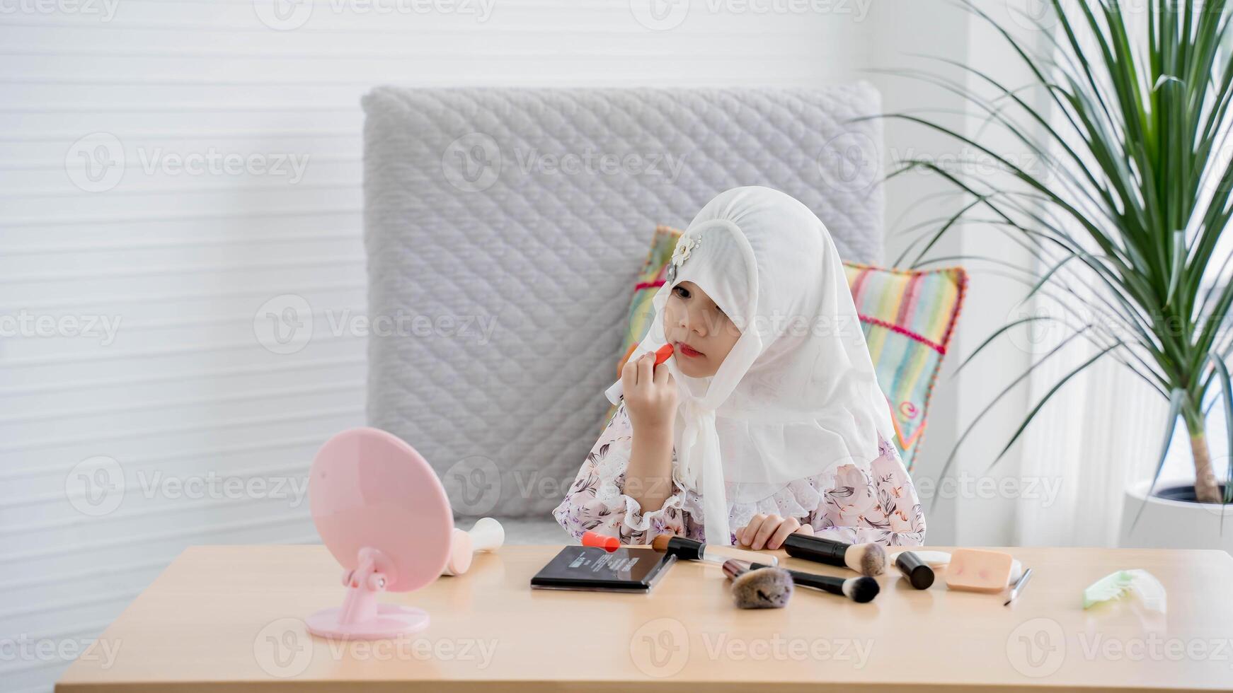 Adorable little girl wearing hijab applying lipstick while looking in the mirror, enjoying her beauty routine surrounded by makeup tools at home. photo