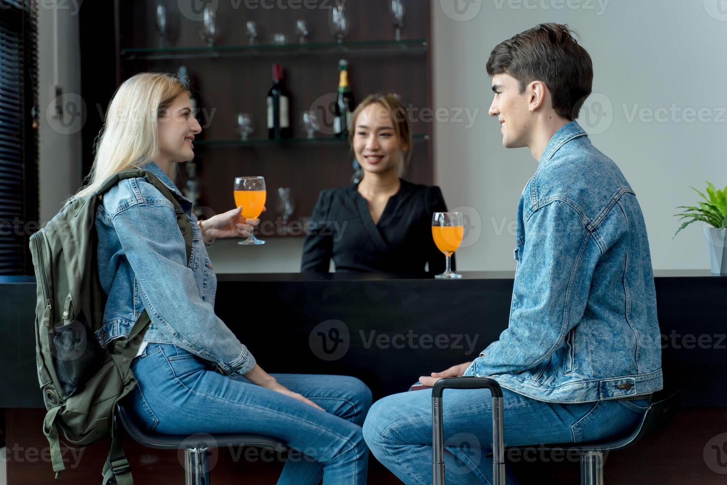 Young couple in denim outfits enjoying drinks at hotel bar counter, smiling and chatting with female bartender during relaxed travel moment indoors photo