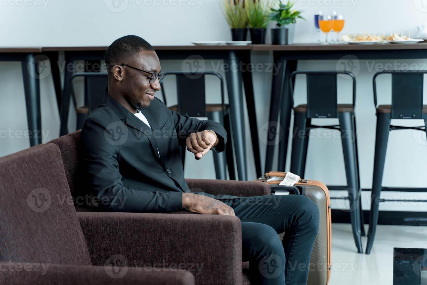 Businessman in suit checking time on wristwatch while waiting with suitcase in modern lounge, travel preparation and punctuality in professional setting photo