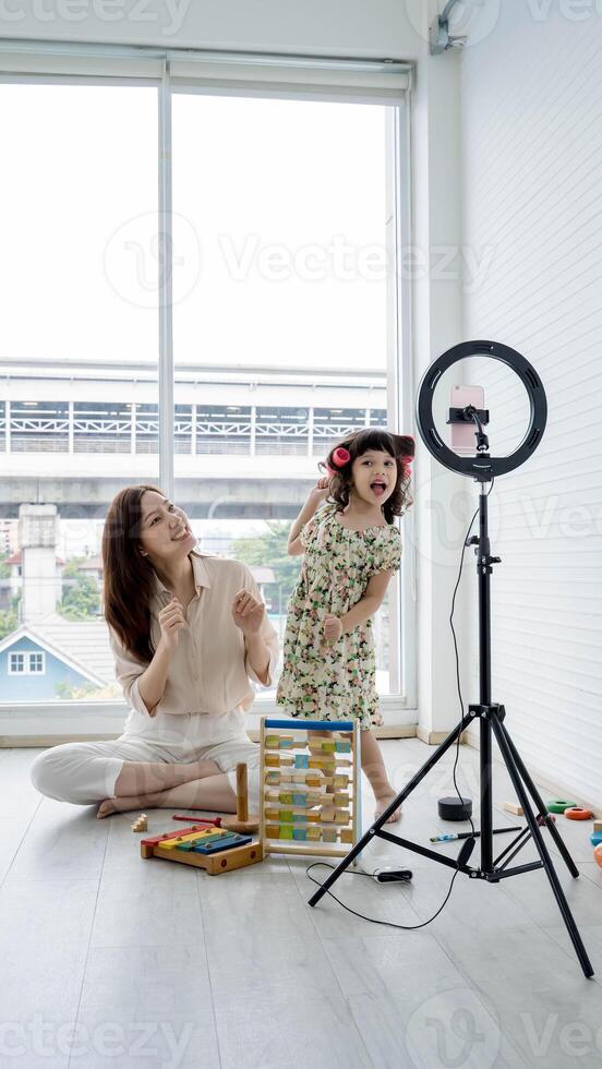 Mother and daughter having fun recording a together at home, smiling and dancing in front of a smartphone on tripod with toys scattered around. photo