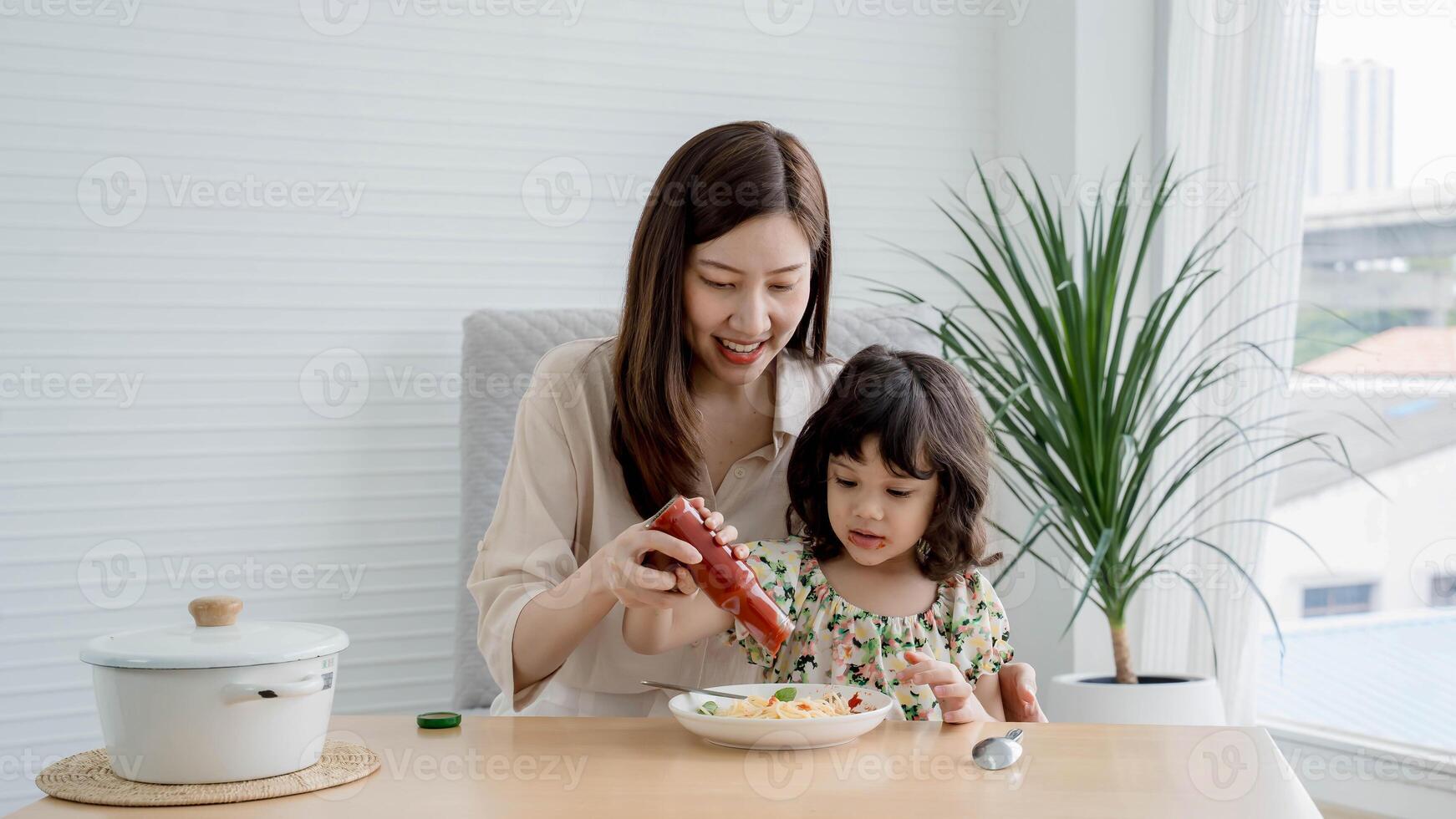 Happy mother and daughter adding ketchup to spaghetti together at dining table, sharing a fun and joyful moment during a home-cooked meal in a cozy kitchen. photo